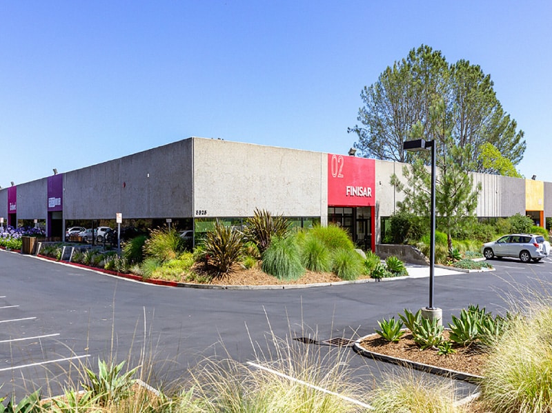 A parking lot view of a small R&D building circled by landscaping